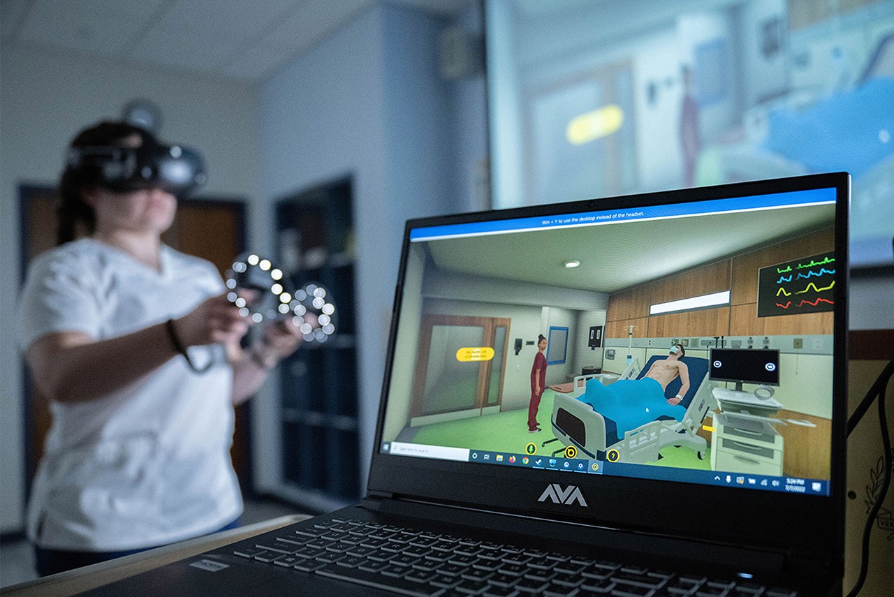 a nurse who's wearing a virtual reality headset and holding controllers in the background stands near a laptop displaying a simulated hospital room 