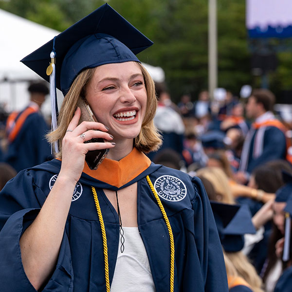 a female Villanova graduate in her cap and gown smiles while on her cellphone