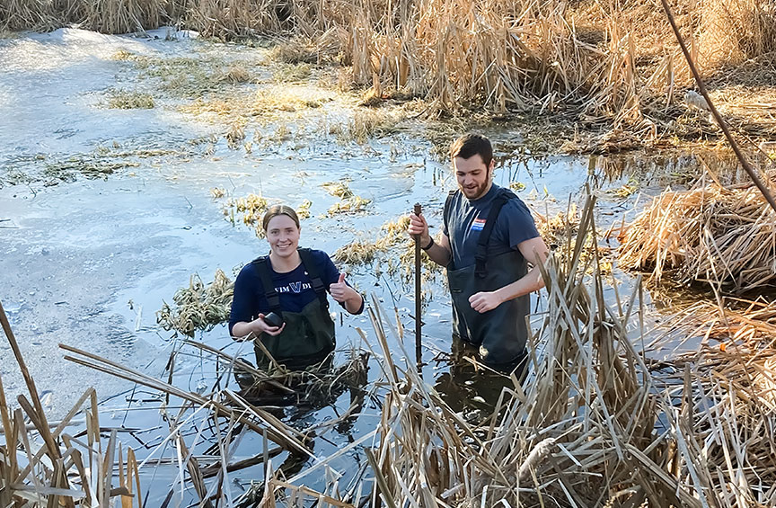 two students wearing waterproof bib overalls standing waist-deep in the wetland on Villanova's campus
