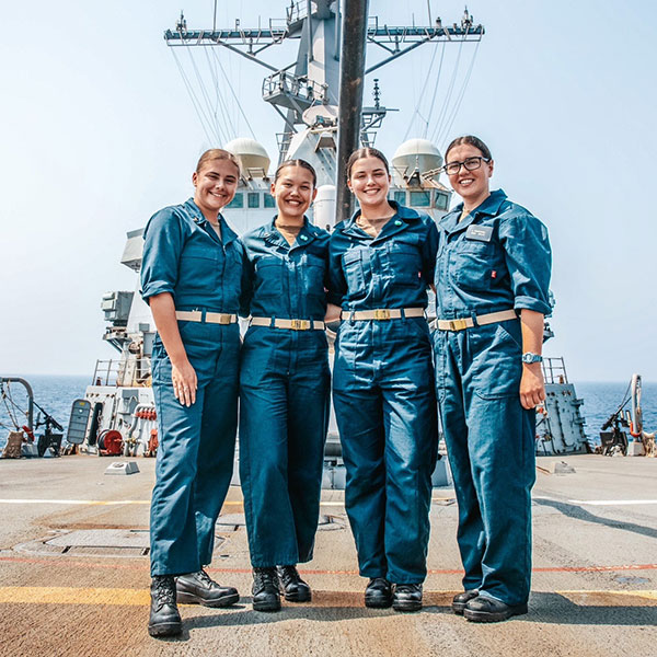 four female members of the NROTC pose in front of a large ship