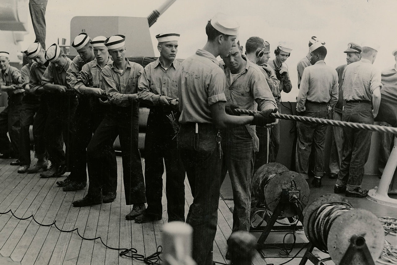 Midshipmen dressed in seaman uniforms heave on a line aboard a battleship in the 1940s