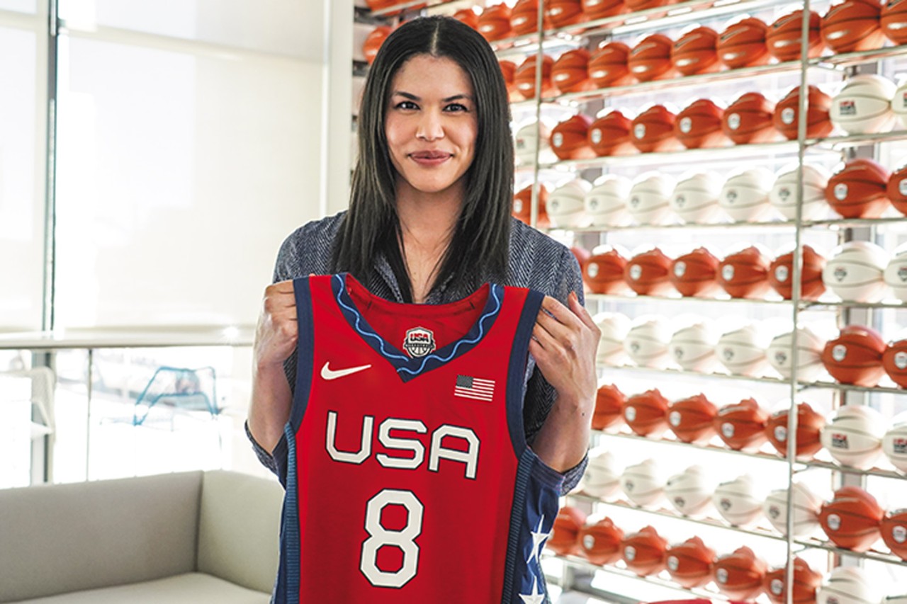 Briana Weiss standing before a wall of basketballs holding up a red USA Women's National Basketball Team jersey