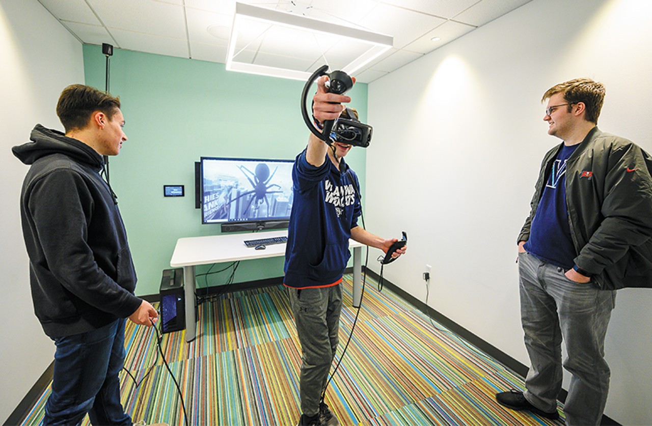 two young men watching a friend playing a virtual reality video game