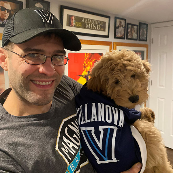 a man wearing a Villanova cap holds a goldendoodle wearing a Villanova dog jersey