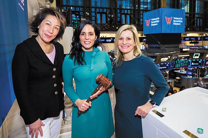 Arianne Collette holds a large wodden gavel as she stands in between two colleagues on the floor of the New York Stock Exchange