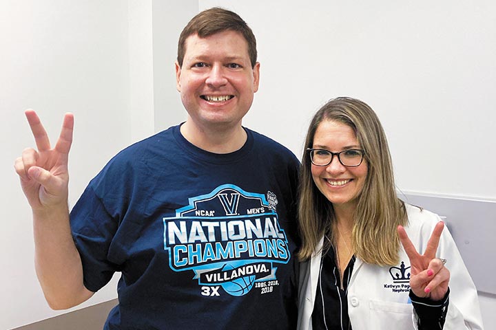 A man wearing a blue Villanova National Chmpions t-shirt stands next to a nurse and they both make the V's up sign