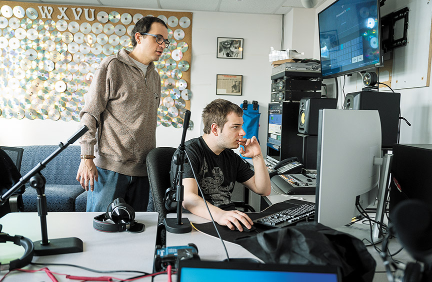 Nick Langan stands behind a seated student who's looking at a monitor in the WXVU studio