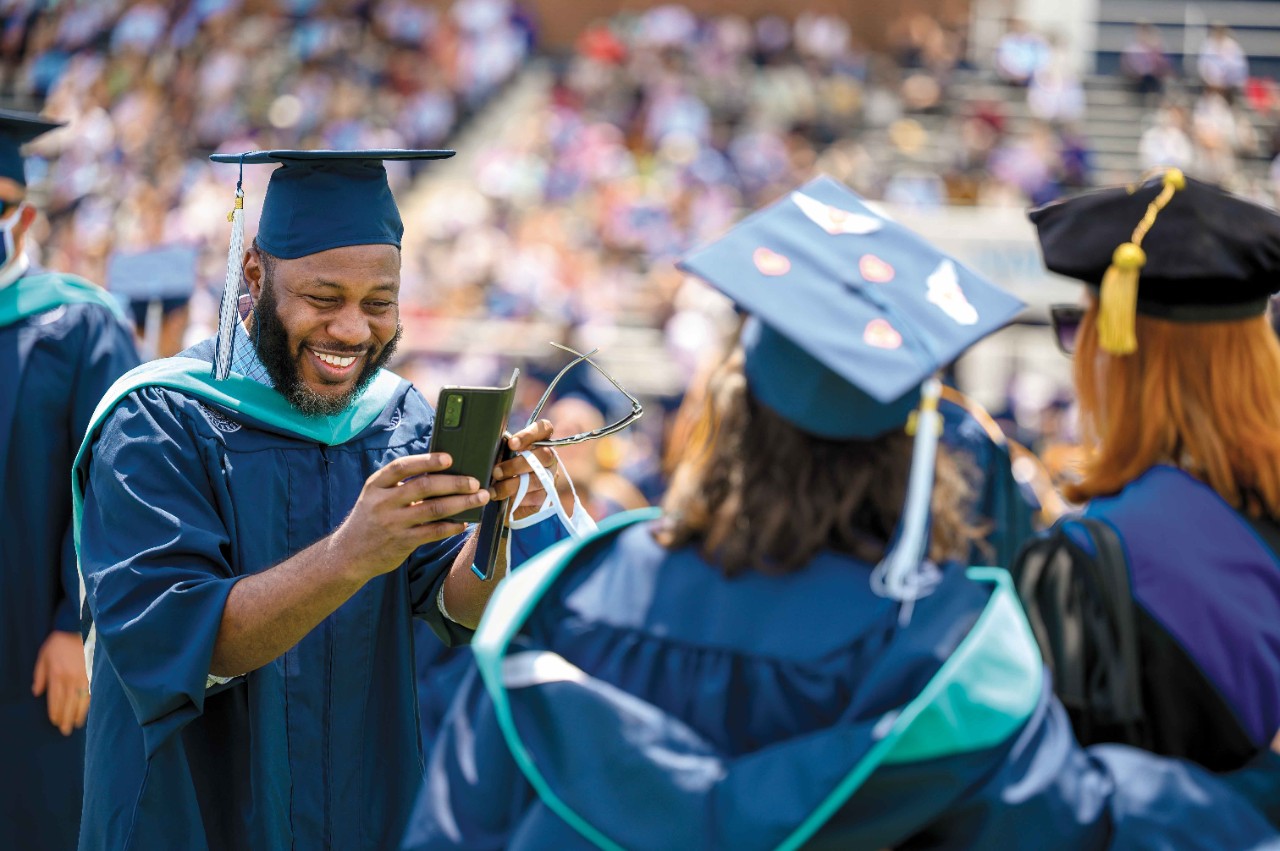 a smiling man in a cap and gown takes a photo of a graduate