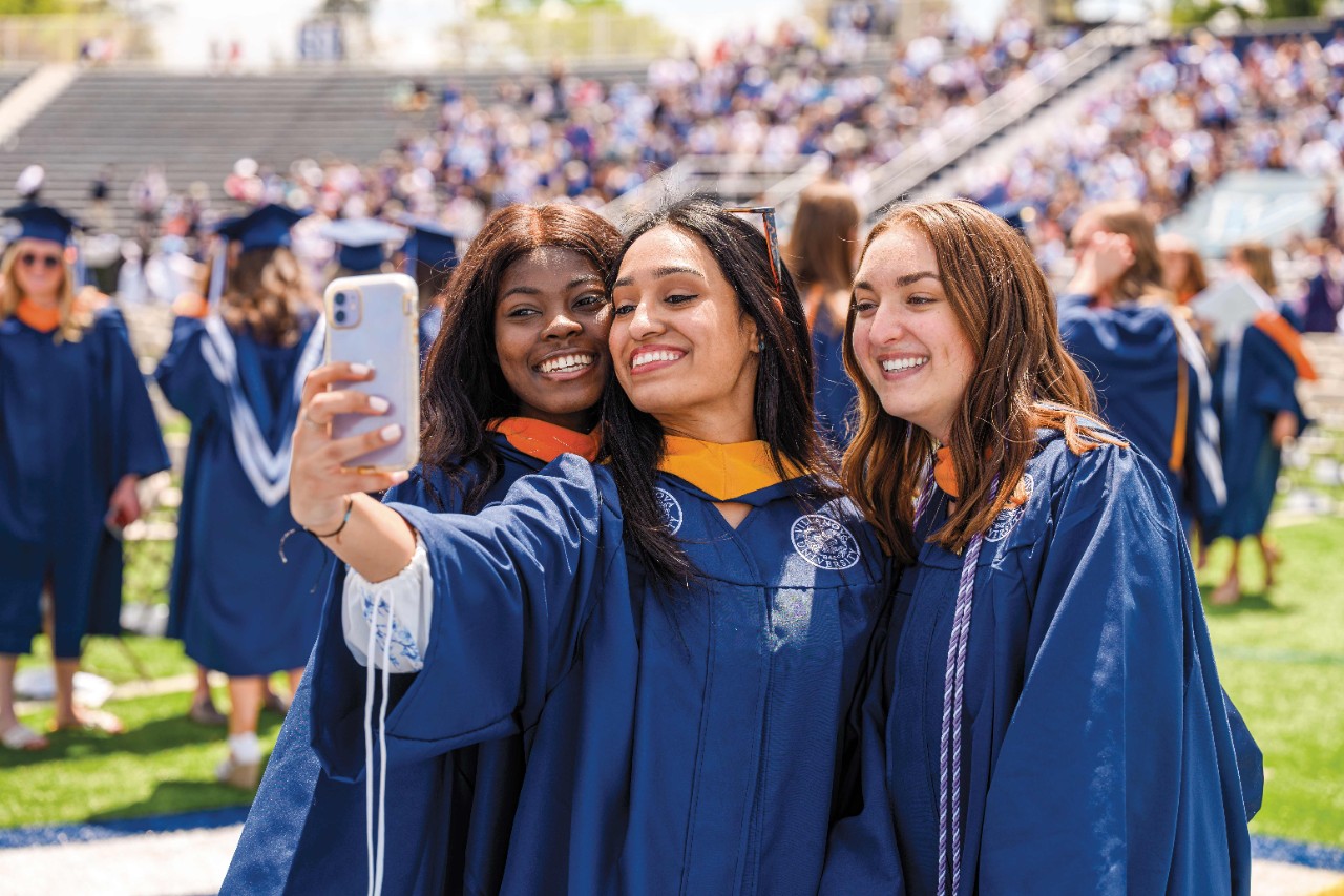 three smiling women wearing blue graduation gowns pose for a selfie at commencement