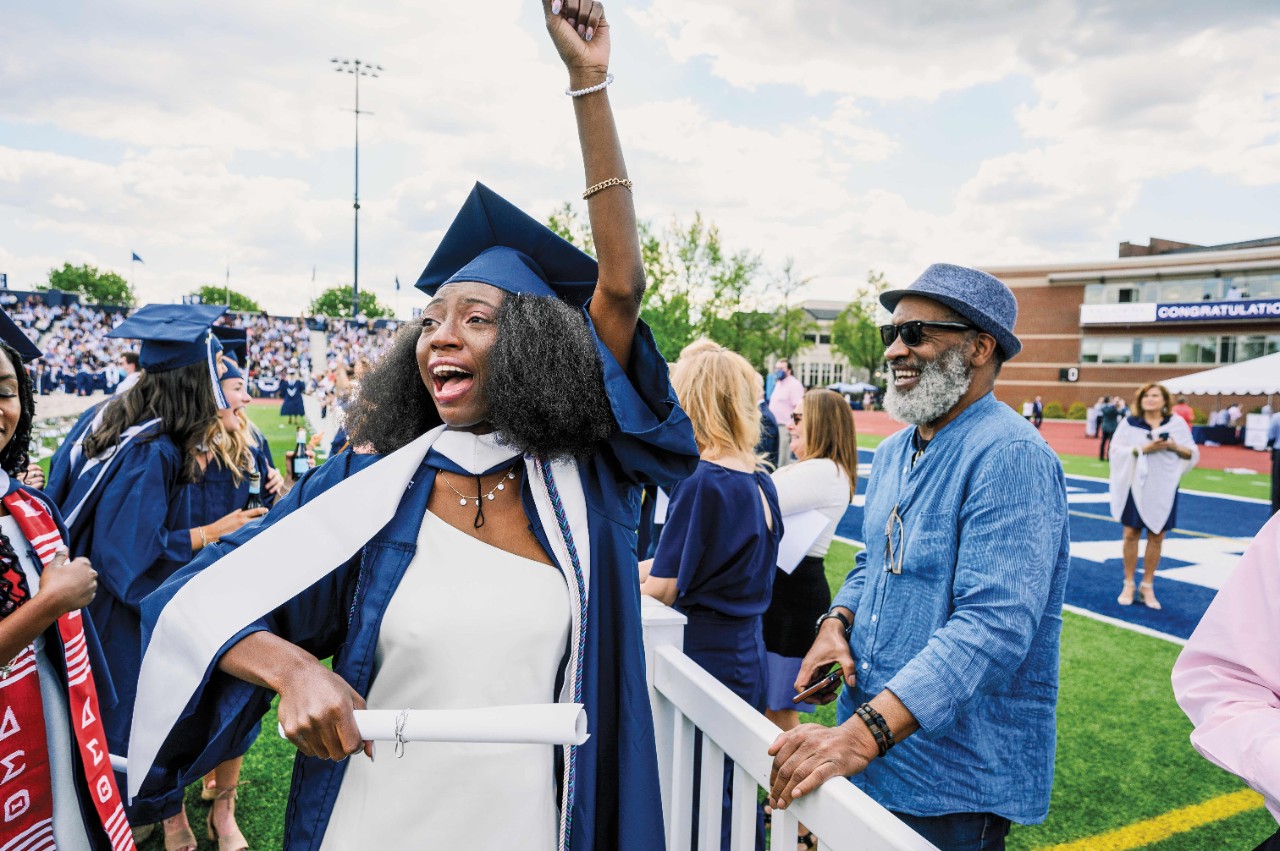 a woman in a blue cap and gown holds up her arm in triumph at commencement