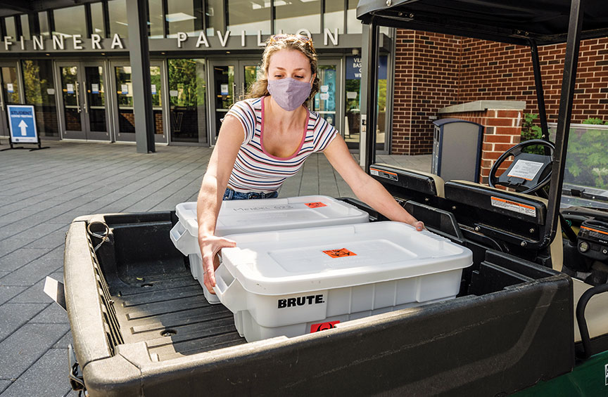 a woman wearing a face mask transports containers with saliva samples outside of Finneran Pavilion