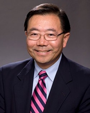 Photo of Stewart Kwoh from the shoulders up. He is smiling and wearing glasses and navy blue suit jacket with a pink tie.