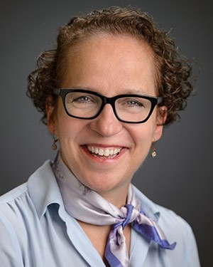 A photo of Dr. Judy Giesberg from the shoulders up. Judy wears a blue button down shirt with a jaunty purple silk scarf tied around her neck. She is smiling.