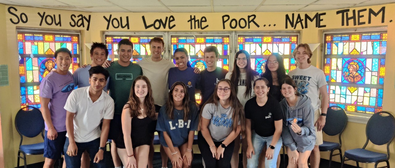 Volunteer students in front of stained glass, wall reads: "So you say you love the poor... Name them."