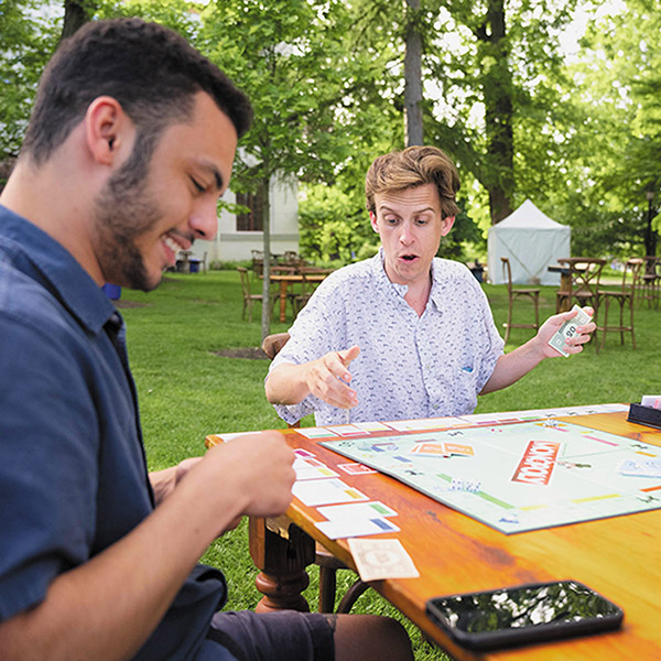 two male students sit outside at a table playing the board game Monopoly